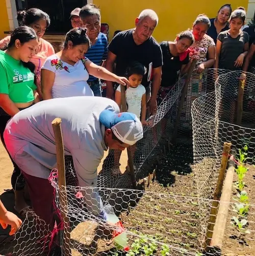 Community members observing a demonstration in a small fenced garden with young vegetable plants growing in neat rows.